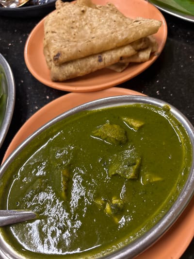 The image shows two dishes of food. On the right, there is a metal bowl filled with a green, thick curry, likely Palak Paneer, an Indian dish made from spinach and cheese. A spoon rests in the curry. To the left, there is an oval orange plate with several rolled flatbreads, possibly roti or chapati. The background is a dark, speckled surface. The scene is likely a restaurant or home meal setting in Kamalapura, India, based on the style of the cuisine and the cuisine type.