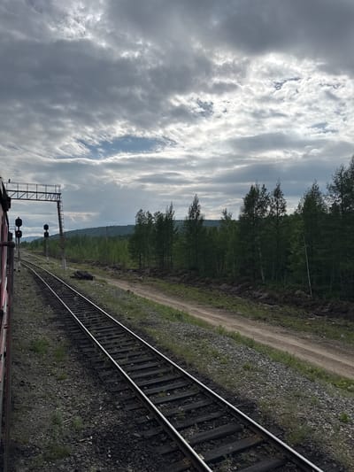 The image captures a view from a train moving along a railway track in Vostochnyy, Russia. The sky is overcast with dramatic, thick clouds, punctuated by streaks of sunlight breaking through in the upper portion of the frame. To the left, the red side of the train car is visible, with a train signal gantry and lights positioned above the tracks. The railway tracks stretch into the distance, bordered by gravel and sparse vegetation. A dirt road runs parallel to the tracks further to the right. Beyond the road and tracks lies a dense forest of coniferous and deciduous trees, with a line of hills forming the horizon. The overall impression is one of a vast, natural landscape under a dynamic sky.