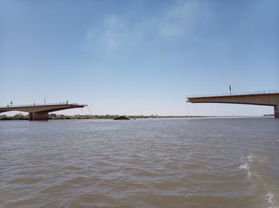 The image shows the collapsed Al-Shambat Bridge in Bahri, Sudan, with two sections of the bridge standing on either side of the river. The central span of the bridge is missing, submerged in the river. The river is brown, and the sky is clear and blue with some clouds. Buildings are visible on the banks of the river in the distance.