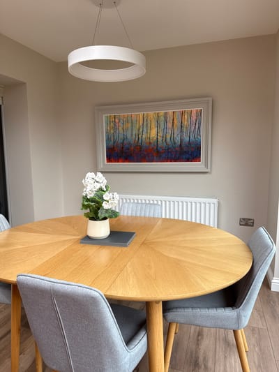 This image captures a modern and neatly arranged dining area, likely within a home. The scene is dominated by a round, light-toned wooden dining table, which features a distinctive radial pattern on its surface. Around the table are three grey fabric upholstered chairs with matching wooden legs, two of which are clearly visible, showcasing their contemporary design.

Above the table, a minimalist white circular pendant light fixture hangs, suspended by thin wires, providing illumination to the space. On the wall opposite the table, a vibrant abstract painting is prominently displayed. The artwork depicts a stylized forest with slender, dark tree trunks set against a lively backdrop of blues, reds, oranges, and yellows, framed in a simple light grey border.

Below the painting and to the right, a white panel radiator is affixed to the wall. Further right and higher up, a silver-colored electrical outlet is visible. On the dining table, a small cream-colored pot containing artificial white flowers, possibly geraniums, sits on a dark grey mat, serving as a centerpiece. The floor throughout the room is covered with light to medium-toned wooden planks, contributing to the warm and inviting aesthetic. The walls are painted a uniform light beige or cream color. No human activity is depicted, and there is no visible text. The overall impression is one of a clean, contemporary, and well-lit dining space.