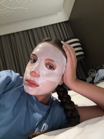 A person wearing a sheet face mask poses for a selfie. The individual, clad in a blue t-shirt, has a long, dark braid. They are resting their hand on their forehead. Behind the person, a striped cushion and wall can be seen. A white sheet is partially visible in the top right corner of the image. The photo has been rotated slightly.