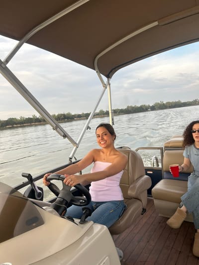 Two women are on a boat. One woman is steering, smiling, wearing a strapless light pink top and jeans. She has dark hair pulled back. The other woman is sitting in the back, wearing a blue shirt and jeans, holding a red cup. She has curly hair and sunglasses. The boat has a canopy and tan seats. The water is calm and there are trees on the shoreline. The sky is overcast.