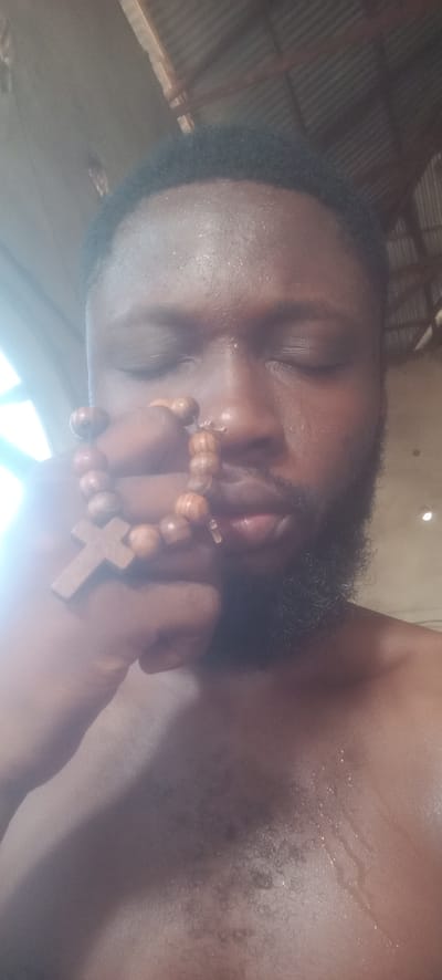 A man with dark skin, closed eyes, and a beard holds a wooden rosary. His skin appears damp, and he is shirtless. The background suggests an interior location, possibly in Bende, Nigeria, based on the request, with a corrugated metal roof visible.