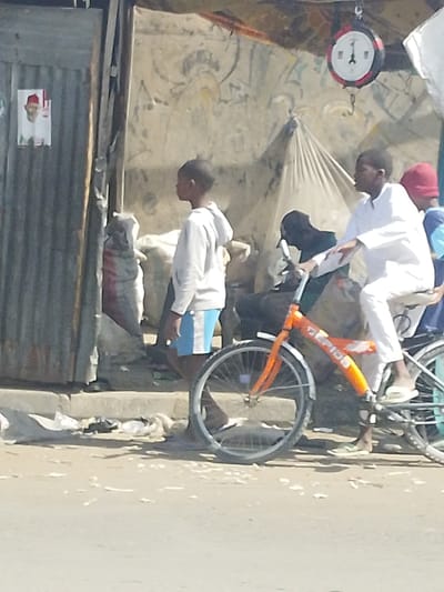 The scene, located in Darmanawa, Nigeria, depicts an outdoor urban environment under daylight conditions. Four individuals, appearing to be children or young adults, are present. In the left foreground, a young male, light-skinned, wears a light grey hooded sweatshirt and blue and white striped shorts; he stands barefoot, facing right in profile. To his right, a second young male, dark-skinned, wears a white long-sleeved garment and white slip-on sandals while seated on an orange bicycle with "ZEIDE" visible on its frame; he holds the handlebars. Behind this individual, a third person, dark-skinned and wearing a dark garment, is partially obscured. Further right, a fourth individual, wearing a red cap and blue garment, is partially visible.
The background features a weathered corrugated metal structure on the left, displaying a poster of a man in a red cap. Adjacent to this, a light-colored, possibly painted, wall with abstract patterns is visible, partially covered by a light-colored canvas or tarpaulin structure. Several large sacks are present near the structures. A red and white circular weighing scale hangs from an overhead support in the upper right quadrant. The ground in the foreground consists of a dusty, light-colored surface with scattered white and light-colored debris near a concrete curb.
