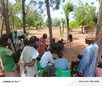 The image depicts a gathering of approximately 20 people, primarily men, in what appears to be a rural or semi-rural area, possibly within Langtang, Nigeria. The scene is predominantly outdoors, with a dirt ground surface and scattered trees providing partial shade. A building is visible in the background.
Several men are standing in a group on the left side of the frame. Others are seated on the ground or on plastic chairs. Clothing styles vary, with some individuals wearing traditional patterned fabrics, while others are in more casual shirts and pants. One man in the center wears a bright orange shirt, standing and seemingly addressing the group. Several people wear hats or head coverings.
A single goat is visible in the background.