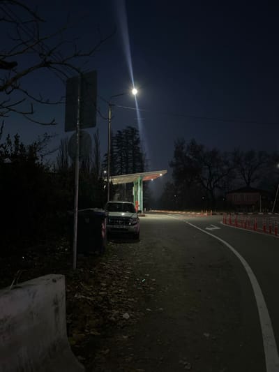 The image depicts a nighttime roadside scene in ლესის თემი, Georgia. The foreground on the left features a concrete barrier, followed by a patch of ground covered in fallen leaves and dark vegetation. A black municipal waste bin is positioned next to a vertical pole displaying two obscured signs. Above, bare tree branches extend into the dark sky.
A street light, mounted on a utility pole, is centrally located, casting a prominent beam of light upwards and illuminating the immediate area with a cool white glow. Directly below the light, a white Ford Escape SUV (Georgian license plate BB-000-XX) is parked beneath a structure with a gray canopy and red illuminated accents, consistent with a fuel station or a checkpoint.
To the right, an asphalt road curves away from the viewer, marked by a solid white line on its right edge and a white arrow painted on the surface indicating a right turn. A series of orange and white vertical traffic bollards or cones delineates the roadside margin. In the background, numerous trees, some deciduous and bare, others coniferous, stand against the dark sky. A single building with a red roof is faintly visible in the far right background, partially obscured by trees. No individuals are present, and no actions or interactions are occurring. The environment is characterized by artificial light against a dark, possibly hazy night.