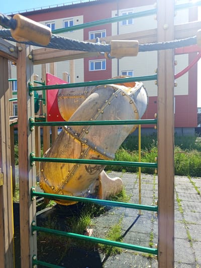 The image depicts a playground slide structure, made of yellow and silver metal, with green horizontal bars in the foreground. A dark rope with yellow cylindrical attachments is stretched across the top of the frame. In the background, a modern apartment building with red and cream-colored siding and white-framed windows is visible. The ground is covered with gray rubberized tiles, interspersed with grass and weeds. The scene suggests an outdoor setting in an urban or suburban area. This scene is located in the city of Gdov, Russia.