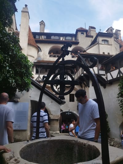 The photograph captures a courtyard within Bran Castle, located in Predeluț, Romania. In the foreground, a large, stone well is visible, with its rough-hewn rim dominating the bottom of the frame. A black metal structure, possibly part of a well mechanism, stands behind the well, featuring ornate scrollwork and a cross-like design.
Several individuals are present in the scene. On the left, a man wearing a grey t-shirt is seen from behind, looking towards a signpost. To his right, a man in a blue and white striped shirt is also facing away from the camera. In the center-right of the image, a man wearing a white t-shirt and sunglasses is leaning over, appearing to interact with the well or examine something near it. In the background, other tourists can be seen on balconies and within the castle's architecture.
The castle itself features creamy-colored stone walls, red-tiled roofs, and numerous arched windows and balconies. Green foliage from trees and ivy is interspersed with the stone structures. The sky above is blue with some scattered clouds. The overall impression is one of historical tourism in an iconic Romanian landmark.