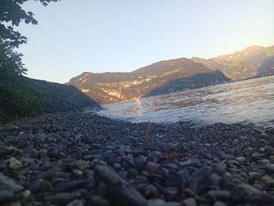 This is a ground-level, wide-angle photograph taken at the edge of a lake, likely Lake Lucerne, in the vicinity of Hergiswil, Switzerland. The foreground is dominated by a dark, pebble-strewn shoreline. Smooth, grey and black stones of varying sizes fill the lower portion of the frame, with some appearing wet from the receding water. To the left, a dark, rough-textured embankment is covered in lush green foliage and tree branches.

The middle ground features the calm, rippling surface of the lake. The water reflects the clear, pale blue sky and the warm, golden light of the setting sun, creating shimmering highlights. In the distance, a range of forested mountains rises steeply from the opposite shore. Several small villages, with buildings clustered together and illuminated by the sunlight, are visible on the lower slopes of these mountains. The peaks of the mountains are illuminated by the sun, revealing their rugged textures and lighter colored rock formations. The sky is clear and mostly devoid of clouds, transitioning from a pale blue at the top to a warmer, yellowish hue near the horizon, indicative of dusk. The overall atmosphere is serene and natural.