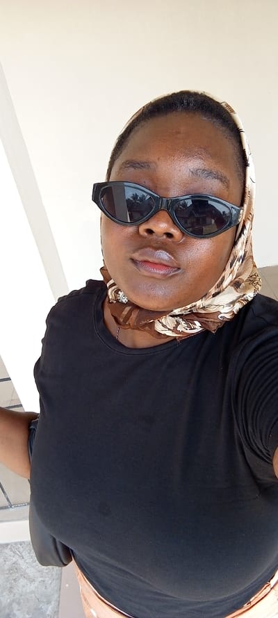 An individual, appearing to be a young adult female with dark skin, is depicted in a close-up, chest-up portrait. She wears black cat-eye sunglasses that obscure her eyes, a plain black crew-neck t-shirt, and a patterned headscarf that covers her hair and wraps around her neck. The headscarf features beige, brown, and off-white tones with an intricate pattern. A faint necklace is visible beneath the scarf. The lower part of a light brown or tan garment is visible at the bottom of the frame. A black bag strap is visible over her left shoulder. The background consists primarily of a light-colored, possibly off-white, wall or structure. To the left, a vertical white architectural element is present. Reflections in the sunglasses show green foliage and what appears to be a building, indicating an outdoor or semi-outdoor setting with natural light. The visible ground at the bottom left is a light grey, possibly concrete or tiled surface. The scene is located in the city of Ijotan, Nigeria.