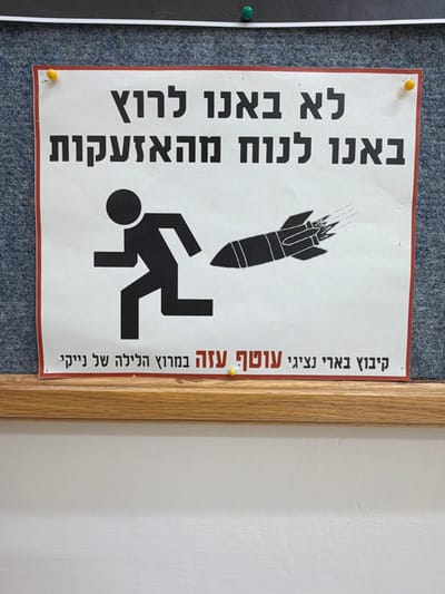 The image features a poster in Hebrew. It displays a graphic of a falling bomb above a stick figure in motion. The text translates to: "We didn't come to run, we came to rest from the sirens." Below this message, it reads: "Kibbutz Be'eri, representatives of the Gaza Envelope in the Nike night race." The poster is pinned to a gray felt board with a wood frame. The background includes a white wall.