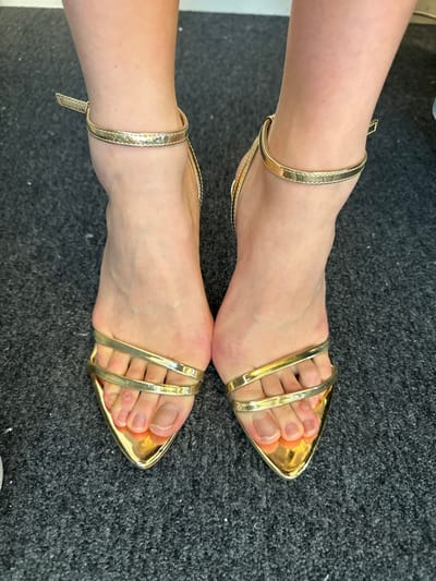 A close-up, top-down view captures a pair of human feet, presumably female, standing side-by-side on a dark gray, short-pile carpet. The scene is located indoors in Arcueil, France. Both feet are adorned with golden, pointed-toe, open-toed high-heeled sandals. Each sandal features a thin strap secured around the ankle and two parallel straps across the top of the forefoot, exposing the toes. The skin tone of the feet is fair. The toenails are visible and appear natural, unpolished, or with a clear coat; the great toenail on the left foot (viewer's left) exhibits a slightly thicker or discolored appearance. The carpet shows a consistent texture across the visible area. A light-colored, possibly white, wall or baseboard is faintly visible at the very top edge of the frame.
