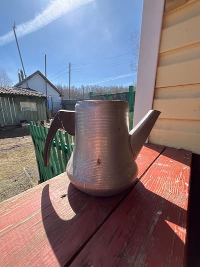 A tarnished metal pitcher with a handle and spout sits on a weathered wooden surface painted red in an outdoor setting. The scene includes a green picket fence, a building with a gray roof, and clear blue sky with a contrail. The location is the city of Верхняя Уря, Russia.