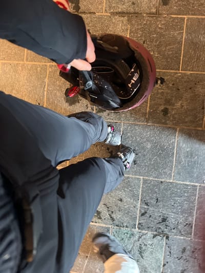 The image depicts a top-down view of a person standing on a tiled ground surface in Valtournenche, Italy, during low-light conditions. The person is wearing black ski pants and grey ski boots with visible buckles and pink accents. A maroon ski helmet rests on the ground to the right of the person's legs. Inside the helmet, a pair of ski goggles with a white strap featuring the "HEAD" brand logo is visible. A red strap or lanyard, partially obscured, appears to have the text "HESTRA" and is attached near the helmet or goggles. A hand, clothed in a dark sleeve, is holding a black object, possibly a ski glove or small bag, positioned near the helmet's opening. The grey tiled ground exhibits several dark, irregular spots, consistent with water or dampness.