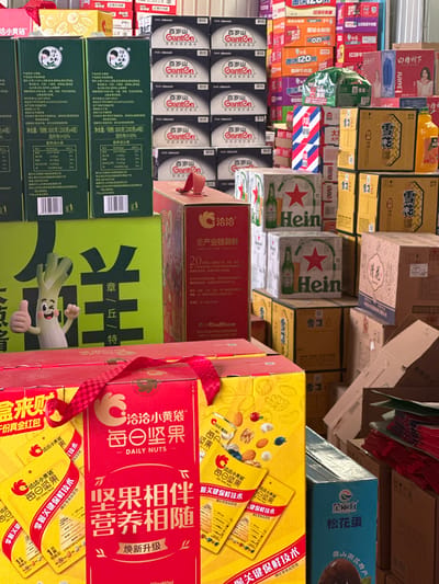The image depicts a crowded commercial storage space in Licheng District, China, filled with stacked boxes of various packaged consumer goods. The foreground is dominated by a large red and yellow gift box of "ChaCha Daily Nuts" (洽洽小黄袋 - 每日坚果), secured with a red checkered ribbon handle and featuring images of mixed nuts. Adjacent to it, a large green box displays a cartoon leek character giving a thumbs-up. Directly behind these, a red box with "ChaCha" (洽洽) branding, labeled "全产业链新鲜," is visible.

In the mid-ground, multiple cases of Heineken beer, identifiable by their green bottles and red star logos, are stacked. To their right, numerous brown and yellow boxes, some possibly for "Snow beer" (雪花), are present alongside a distinct vertically striped red, white, and blue box. The background features tall, dense stacks of black and white boxes labeled "Ganten" (百岁山) mineral water. Other colorful boxes, including orange, pink, and blue, display various food and beverage items, with visible text such as "O2" and "JUICE," and one box showing a woman's image. The overall scene indicates a high volume of goods in an indoor commercial environment. No individuals are visible.
