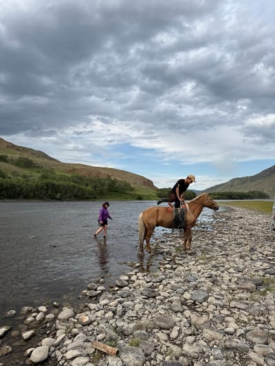 An outdoor scene in Burentogtoh, Mongolia, features a river flowing through a valley under a partially overcast sky. The sky transitions from dark, heavy clouds on the upper left to lighter grey and patches of blue on the upper right. Rolling green hills line both sides of the wide river in the background.
The foreground consists of a rocky riverbank composed of various sizes of rounded, light-colored stones, with some small green vegetation patches and a segment of a light brown pipe or log on the lower left. The river's water appears clear and shallow near the bank, revealing submerged rocks.
Two individuals and one horse are visible. On the left, a person wearing a purple long-sleeved top, black shorts, and a dark cap stands in the river's shallow water, with water reaching their shins, appearing to wade deeper. On the right, a light brown horse, equipped with a dark saddle and bridle, stands on the rocky bank at the river's edge. A second person, wearing a dark short-sleeved shirt, dark pants, and a light-colored hat, is positioned on the horse's back, mid-action, with their left leg lifted high and bent over the horse, suggesting mounting or dismounting. A partial view of a light-colored vehicle is visible on the far right edge of the frame, parked on the rocky bank.