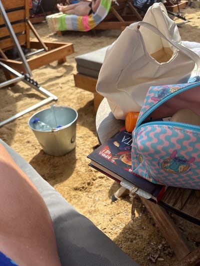 The image depicts a close-up view of a beach scene in Nea Iraklitsa, Greece, focusing on items placed on a wooden table. In the foreground, a person's leg is visible, resting on a grey fabric surface, likely a beach chair. To the right of the leg, a silver metal bucket containing two plastic water bottles is partially visible, resting on the sandy ground. On the wooden table, a white tote bag is laid open. Next to it, a small, pink and blue patterned zippered pouch featuring cartoon characters is also open, revealing some contents inside. A book with a dark cover and an orange cap object are also on the table. In the background, beach chairs and people relaxing on the beach can be seen, with the general environment suggesting a sunny day at the coast.