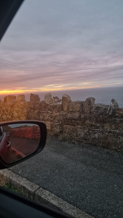 The image captures a coastal view from a car, likely on the Great Orme in Llandudno, United Kingdom, during a sunset. A rough stone wall, characteristic of the area, runs horizontally across the frame, with the sea visible beyond. In the reflection of the car's side mirror, a stone wall is visible along a road. The sky is a canvas of soft pastel colors, with streaks of orange and pink from the setting sun. A sheep is perched atop the stone wall, gazing towards the sea. The ground in front of the wall is covered in dark gravel.