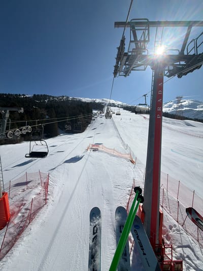 The image presents a first-person perspective from a ski lift chair overlooking a snow-covered ski slope in Valdisotto, Italy, under a clear blue sky. In the immediate foreground, the tips of two skis, identified as "HEAD SHAPE JZ," and two bright green ski poles are visible, angled downwards.

The main subject is a wide ski slope covered in groomed snow, descending into the distance. The slope is delineated by red and orange safety netting on both sides. Numerous ski lift chairs are visible along two parallel lines, ascending the mountain. Some chairs are empty, while others contain individuals. Shadows of ascending ski lift chairs are cast prominently on the pristine snow.

To the right, a gray ski lift tower, labeled "4" and vertically branded with "PRADA" in red lettering, rises towards the upper right corner. The sun is positioned behind and to the left of this tower, casting a strong starburst glare. To the left of the slope, a dense line of coniferous trees marks the edge of the prepared run, giving way to further snow-covered mountain terrain. In the upper background, additional snow-capped peaks are visible, and more ski lift infrastructure extends into the distance. Several individual skiers are discernible descending the slope at various points. The overall environment is mountainous, characterized by extensive snow cover and clear weather.