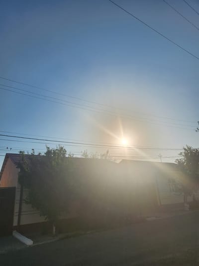 The image depicts a clear, bright sky over a residential area. The sun is visible, casting a strong glare and lens flare across the scene. Several power lines are strung across the sky. In the foreground, partially obscured by the sun's glare and shadows, are the roofs of houses, trees, and a paved road. The architecture suggests a typical suburban or rural setting. The overall lighting indicates it is daytime, likely morning or late afternoon, given the low angle of the sun. The image does not contain any discernible individuals or explicit actions.