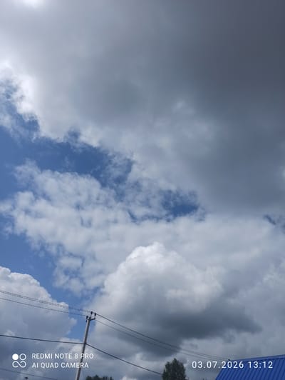 This is a landscape photograph taken outdoors in Sovetskiy, Russia, on July 3, 2026, at 13:12. The image is dominated by a sky filled with a mix of blue sky and dramatic clouds, ranging from bright white to dark gray. A utility pole with power lines is visible in the lower half of the frame, extending upwards towards the sky. The bottom edge of the image shows a glimpse of what appears to be a blue corrugated roof of a building and the tops of some trees. A watermark in the lower left corner reads "REDMI NOTE 8 PRO AI QUAD CAMERA," and the date and time stamp "03.07.2026 13:12" is located in the lower right corner. No people are visible in the image.