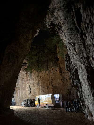 The image shows the interior of a cave, likely located near Guttapadu, India, as suggested by the context. The cave entrance is wide, with rough, textured rock formations on either side creating a natural frame. The lighting is dim, with a spotlight illuminating the center of the cave floor.

In the mid-ground, two vehicles are present. On the left, a dark-colored van or SUV is parked, with its rear door open and several individuals gathered around it. They appear to be engaged in some activity, possibly loading or unloading equipment. To the right of this vehicle is a yellow and white bus or small truck, also parked.

Further to the right, a group of tripods, likely for cameras or lighting equipment, are set up, suggesting a film or photo shoot is in progress. Some individuals are visible near this equipment.

The cave walls are rough and striated, displaying earthy tones of brown and gray. Patches of green foliage are visible clinging to the upper parts of the rock faces, indicating the presence of vegetation outside the cave. The ground appears to be made of dirt or loose gravel. The overall atmosphere is that of an operational outdoor setting within a natural geological formation.