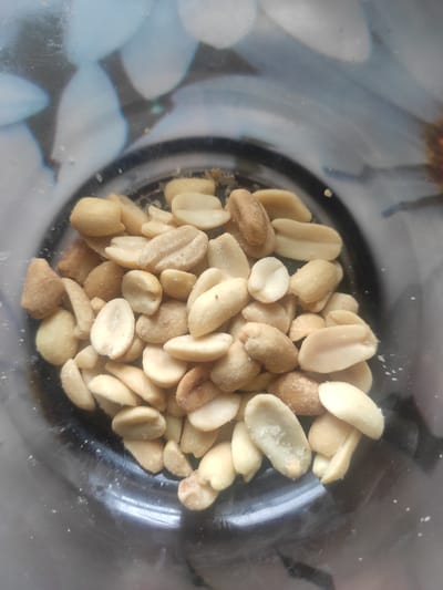 A close-up, top-down shot shows a pile of roasted peanuts inside a clear glass jar. The peanuts are light brown and have a slightly wrinkled texture. Some of them are broken in half, revealing their interior. Small white salt crystals are visible on some of the peanuts. The background inside the jar is blurred, showing a pattern of pale blue and white shapes that appear to be flowers. The exterior of the jar shows reflections of a floral pattern, possibly a tablecloth or wallpaper. The image does not contain any people or evidence of specific locations within the city of Kostopil, Ukraine.