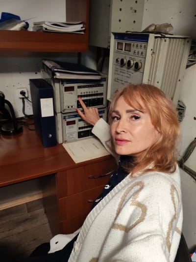 A woman with light auburn hair and makeup is seated at a wooden desk in a room that appears to be a control room or technical office, possibly located in Belousovo, Russia. She is wearing a white cardigan over a dark shirt and is pointing at a control panel with a red-tipped fingernail. The control panel is stacked with two units, featuring buttons, knobs, and digital displays. To the left of the control panel is a dark blue binder labeled "ЭМС" (EMC). On the desk, there is also a microphone, papers, and a stack of files on a shelf above. The wall behind the equipment is white and shows some signs of wear.