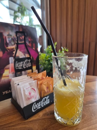 The image presents a close-up view of a half-full glass containing a frothy yellow beverage and a black bent plastic straw, resting on a light brown wooden table. The glass features a geometric diamond-cut pattern on its lower exterior and exhibits visible residue or condensation on its upper interior. To the left of the glass, a black rectangular condiment holder branded with the "Coca-Cola" logo holds white individual packets labeled "Portion" and orange individual packets labeled "Gevgelija," consistent with sugar or sweetener products. Behind the condiment holder, a vertical advertising display board is visible, featuring a dark background with graphics of Coca-Cola bottles and wine glasses, and partially obscured Cyrillic text. A small green artificial plant is situated between the holder and the beverage glass. The background consists of a blurred view through a large glass partition, revealing an indistinct silhouette of a person to the left of the glass, and a vertical wooden slatted wall to the right. This scene is situated within a cafe or restaurant environment in Gevgelija, North Macedonia.