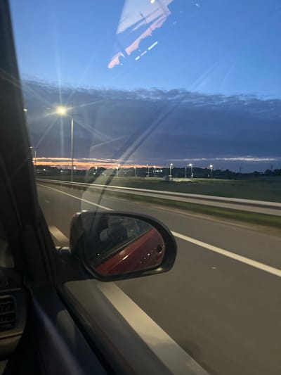 The image is a first-person perspective shot from inside a vehicle, looking out the side window. It captures a view of a highway at dusk. The sky is a deep blue transitioning to orange and pink near the horizon, indicating either sunrise or sunset. Clouds are visible in the upper portion of the sky. Streetlights line the side of the highway, casting a warm glow. The road surface is dark asphalt with a white lane marker. A guardrail separates the highway from a grassy verge. In the foreground, a portion of the car's interior is visible, including the side mirror which reflects the scene. The reflection in the side mirror shows the interior of the car and a person holding a phone. The overall scene depicts travel on a highway during twilight, with an urban or suburban landscape visible in the distance. There is no specific indication of the location being Vasil'kovo, Russia.