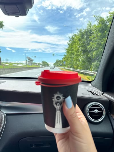 A person's hand with light blue painted fingernails holds a black paper coffee cup with a red lid, in front of a car's dashboard. The cup has a white logo with a sun-like figure. The dashboard is dark leather with silver accents, including an air vent visible to the right. Outside the car, seen through the windshield, is a highway with other vehicles, lush green trees lining the road, and a bright blue sky with scattered white clouds. The image appears to be taken from inside a moving vehicle, suggesting a journey through a suburban or rural area. The city of Ir, Russia, is not identifiable from the visual information in this image.