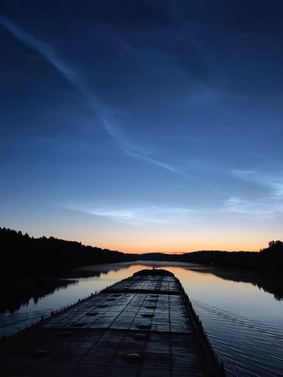The image depicts a view from the deck of a barge on a body of water, likely a river, at dusk or dawn. The sky is a deep blue with wispy, luminous clouds, suggesting noctilucent clouds which are sometimes visible at high latitudes. The horizon is illuminated by a soft orange glow, indicating the sun is low or has recently set/risen. A silhouetted treeline borders the water on both sides, with a faint mist rising from the water's surface in the distance. The barge itself is dark and textured, with visible panels and raised circular objects. Its wake is visible trailing behind it, indicating movement. The overall impression is one of quiet, natural beauty and solitary passage. The location in the city of Volokov Most, Russia, is not explicitly visible or indicated in the image itself.