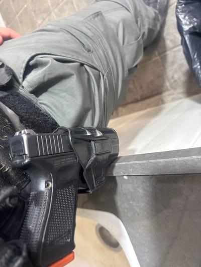 The image presents a high-angle, close-up view of an individual's lower torso and legs within an indoor restroom environment in the city of Lod, Israel. The person is seated, wearing grey tactical or cargo pants. A black semi-automatic pistol, consistent with a Glock model, is holstered on the person's right hip (from the wearer's perspective). The pistol is secured in a black polymer holster, bearing a "G-9" brand mark, which is affixed to a black belt or tactical gear partially covered by a green mesh material. The visible portion of the pistol features a textured grip and an orange baseplate on the magazine. The background elements include light-colored tiled walls, the rim and interior of a white toilet bowl, and a crumpled black plastic bag positioned in the upper right. The person's left hand rests on their left thigh.