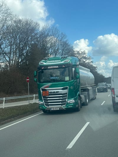 The image displays a daytime traffic scene on an asphalt road, located in or near Kirchdorf am Inn, Germany. The central subject is a dark green DAF semi-truck, registered with Austrian license plate "WL KG 69," displaying "GASPERLMAYR" and "AUSTRIA" branding. The truck pulls a silver, multi-axle cylindrical tanker trailer. An orange hazard placard on the truck's front bumper indicates "30" (flammable liquid) over "1202" (Diesel fuel / Gas oil). The name "AYDIN 24" is visible inside the truck's windshield.

The truck is positioned in what appears to be the left lane of traffic, alongside a white commercial van partially visible in the foreground on the right. A queue of passenger vehicles, including a dark SUV, proceeds ahead in the same lane as the DAF truck. The road features white dashed lane markings. To the left of the roadway, a metal guardrail runs parallel to a grassy verge, beyond which stands a dense line of leafless deciduous trees. A red circular "No Entry" road sign with a white horizontal bar is posted on a pole on the left roadside. The sky is blue with scattered white cumulus clouds. No pedestrians are visible.