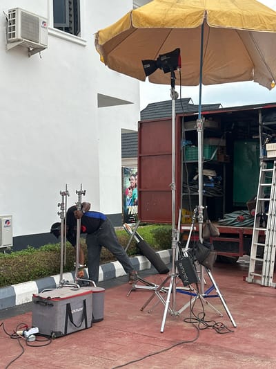 An exterior shot captures a film or photography setup in an urban environment, specifically Pakuro, Nigeria. In the foreground, a man in a dark blue shirt and grey jeans is adjusting two light stands. Next to him are two grey fabric cases, one with wheels, labeled "Aputure." A power strip with electrical cords is plugged into the ground. Several other light stands, some with attached equipment, are positioned around the scene. To the right, a large red truck with its rear doors open reveals shelves stocked with equipment, including coiled cables, crates, and a green generator. A silver ladder leans against the truck. Overhead, a large yellow umbrella, possibly for diffusion, shields some of the equipment. A white building with an air conditioning unit is visible on the left. A large poster featuring actors is displayed near the right edge of the frame. The ground is a reddish-brown paved surface.