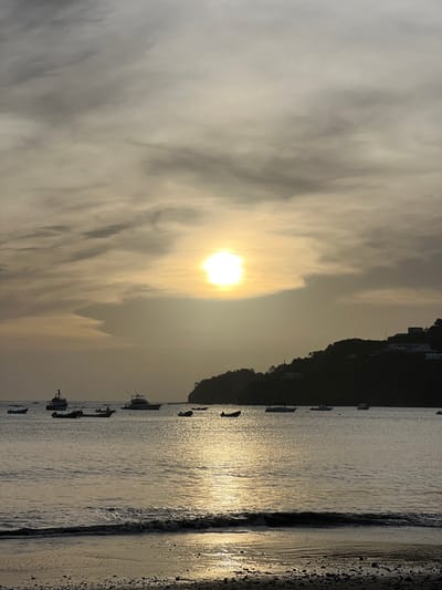 The image depicts a serene sunset scene at the beach in San Juan del Sur, Nicaragua. The sun, partially obscured by clouds, casts a golden glow across the sky and reflects on the calm waters. Several boats are anchored in the bay, their silhouettes contrasting with the luminous sky. A lush, green hill rises in the background, dotted with buildings, suggesting a coastal town. The foreground shows a pebble beach with gentle waves lapping at the shore. The overall atmosphere is tranquil and picturesque, characteristic of a tropical sunset.