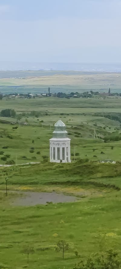 The image shows a distant, white, octagonal structure with a green, multi-tiered roof, set within a vast, rolling green landscape. The structure has tall, narrow windows or columns along its sides. The surrounding environment consists of gently sloping hills covered in grass, with scattered trees and some signs of human habitation in the far distance. A body of water, possibly a pond or small lake, is visible in the foreground, reflecting the overcast sky. The sky itself is a pale, hazy blue, with faint outlines of distant hills and what appears to be a city or settlement on the horizon. There are no people or animals visible in the scene. The image appears to be taken from an elevated vantage point, looking out over the landscape towards the distant horizon. The location is identified as Nigatun, Armenia.