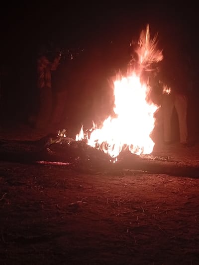 A large bonfire roars in the center, illuminating the dark ground. Silhouetted figures stand around the fire, one seemingly playing a trumpet. Large logs fuel the intense flames. The ground is textured with dirt and scattered debris. The scene is shrouded in darkness, the fire casting a warm, vibrant glow on the surrounding area.