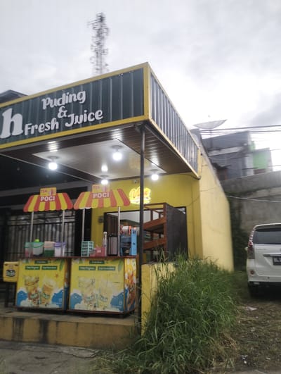 A bright yellow building with a green awning that reads "Puding & Fresh Juice" stands in the foreground. Under the awning, two small kiosks with yellow and red striped awnings display promotional images for "Teh Poci" (Teh Poci tea). The kiosks are filled with various containers and equipment, suggesting a food or beverage establishment. In front of the kiosks, two larger displays showcase images of drinks and text such as "Fresh Brew" and "Tanpa Pengawet Tanpa Pemanis Buatan" (No Preservatives, No Artificial Sweeteners). A white van is parked to the right of the building, and tall, overgrown grass and weeds are visible in the foreground and to the right. The sky is overcast, suggesting a cloudy or potentially rainy day. In the background, a tall cell tower and satellite dish are visible, indicating an urban or semi-urban setting. The overall scene depicts a small business operating outdoors, likely a casual eatery or drink stand. The text "TEH POCI" is prominently displayed on the kiosks.