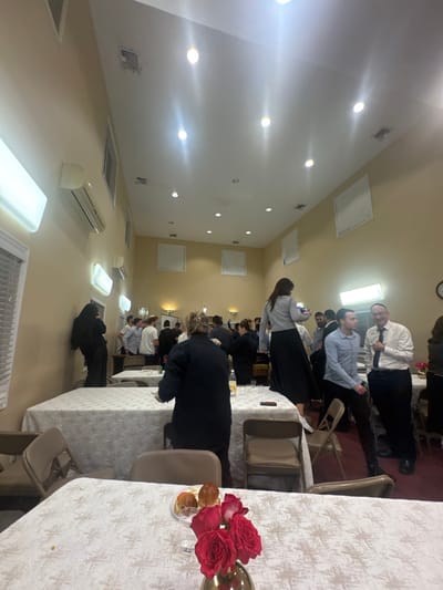 The image depicts an indoor gathering in a large, well-lit hall located in Bergenfield, United States. The hall features light-colored walls, a high vaulted ceiling with numerous recessed spotlights, and several wall-mounted split-unit air conditioning units and rectangular fluorescent light fixtures. Multiple windows with white blinds are visible on the left wall, and square vent covers are present on the ceiling and upper walls.
In the foreground, a table covered with a white, textured tablecloth holds a vase containing three red roses and a small bowl with several pastries.
Mid-ground, a woman in a gray long-sleeved top and a long black skirt is standing on a brown folding chair, holding a mobile phone in her hands, facing away from the viewer. To her right, a man in a light blue button-down shirt and dark pants stands, looking towards the right side of the frame. An older man to his right, wearing a white button-down shirt, dark tie, and kippah, holds a black object, possibly a microphone, and is speaking or gesturing. Another woman in a dark top and skirt is partially visible behind the man in the blue shirt. Near the center-left, a woman in a dark garment stands with her back to the viewer, positioned near a table with a yellow bottle.
In the background and scattered throughout the hall, approximately twenty to thirty individuals are observed standing and interacting. Several men are wearing kippahs. On the far left, a woman in a black garment leans against a wall, looking at a mobile phone. Tables covered with white tablecloths and numerous brown folding chairs, some occupied and some empty, are arranged throughout the space. A wall clock is visible on the far right wall. The scene suggests a social or community event.