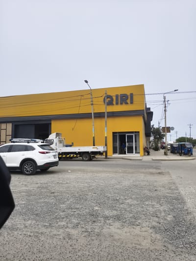 This image depicts a street scene in Nuevo Chimbote, Peru, under an overcast sky. The central feature is a large, brightly yellow-painted building with a dark grey fascia and large, dark openings, one of which appears to be a main entrance with glass doors. The word "QIRI" is prominently displayed in dark grey letters on the upper part of the yellow facade.
In the foreground, a white Mazda CX-9 SUV with roof racks, carrying what appears to be a surfboard or similar item, is positioned on a gravel and dirt surface, facing right. Its license plate, "BF-284," is visible. Behind the SUV and adjacent to the building, a white flatbed truck with "AXO 7US" written on its side is parked.
Two individuals are seated on a low wall or bench to the right of the main entrance of the yellow building. One person in a blue shirt is visible inside the glass entrance. Further down the street to the right, several blue three-wheeled mototaxis are parked.
Numerous utility poles with extensive black wiring crisscross the scene, some supporting streetlights. One pole near the center of the image has yellow and blue paint at its base. The immediate ground consists of gravel and dirt, transitioning to a paved sidewalk directly in front of the yellow building. A small tree is visible on the sidewalk further down the street, and additional faint structures and vehicles are discernible in the distance.