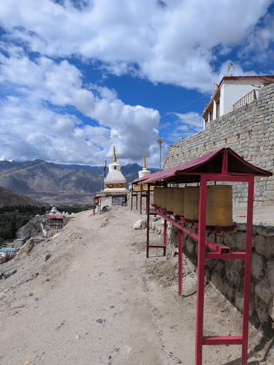 The image captures a Buddhist monastery located in Choma, India, perched on a hillside with a mountainous backdrop. In the foreground, several large golden prayer wheels are mounted on a red metal rack, casting shadows on the sandy ground. A dirt path leads away from the prayer wheels towards a white stupa with gold accents, which is situated on a raised platform. Further in the distance, more stupas and buildings of the monastery complex are visible, nestled into the hillside. The sky above is a vibrant blue, adorned with scattered white clouds. The surrounding environment is arid and rocky, typical of a high-altitude region. No people are visible in the scene.