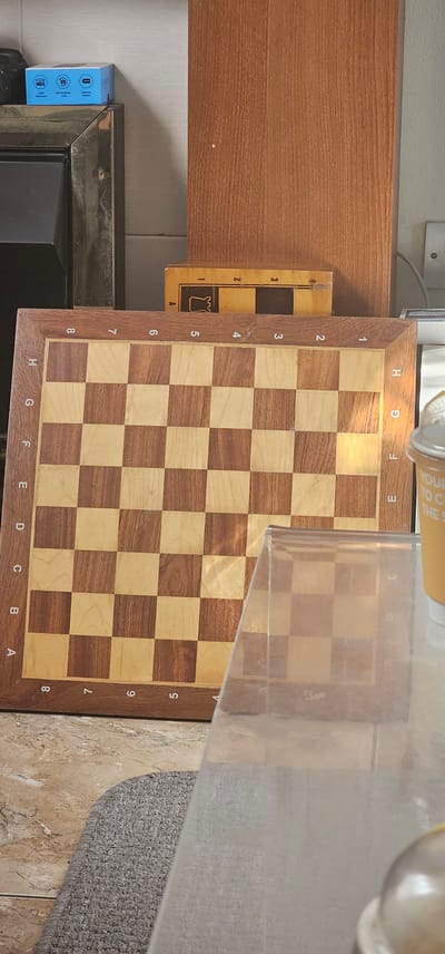 The image shows a chessboard from a top-down perspective, oriented vertically on a wooden surface. The chessboard is made of alternating light and dark wood squares. Numbering from 1 to 8 runs along the left side of the board, and letters from A to H run along the bottom. A clear plastic cover is placed over a portion of the chessboard, obscuring some of the squares. To the left of the chessboard, another smaller yellow board with a black and white checkered pattern and the number 3 and a chess piece icon is visible.
In the background, a wooden shelf runs horizontally across the top of the image, with a light-colored wall behind it. To the left, a blue box with some icons and text is partially visible on a darker surface, which appears to be part of a furniture frame. The lighting suggests an indoor setting, likely during the daytime, with a bright light source casting a warm glow on the chessboard. There are no people visible in the image.
Visible text includes numbers and letters on the chessboard itself, and partial text on the yellow board to the left which reads "3", "2", "1", and "A", along with a chess piece symbol. A partially visible cylindrical object with yellow and white text is at the top right of the image. The text on this object includes "YOUR" and "TO C", and "THE S". The blue box on the left has text and icons, but they are too small to read.