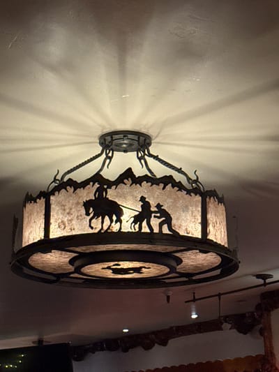 An ornate light fixture with a circular lampshade casts rays onto the wall. The lampshade depicts silhouettes of cowboys and cattle. It is attached to the wall with metal arms and a circular base. Parts of a room are visible on the right, including decorative molding and a dark space.