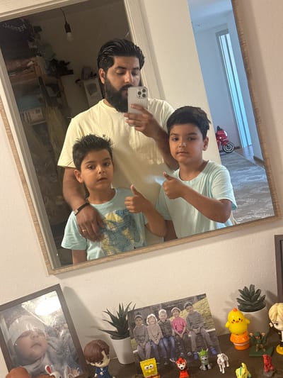 A man is taking a mirror selfie with two young boys in what appears to be a residential interior. The man, with dark hair and a beard, is wearing a light yellow t-shirt and holding a smartphone. The two boys, also with dark hair, are standing in front of him. The boy on the left is wearing a light blue t-shirt with a graphic print and is giving a thumbs-up. The boy on the right is wearing a light blue t-shirt and is also giving a thumbs-up. The background reflected in the mirror shows a doorway, a red toy motorcycle, and a hallway. On a shelf below the mirror, there are several framed photographs and small figurines, including a framed picture of a baby, a group photo of children, and various cartoon character figurines. The overall environment suggests a home setting, possibly in Mainburg, Germany, given the context provided.