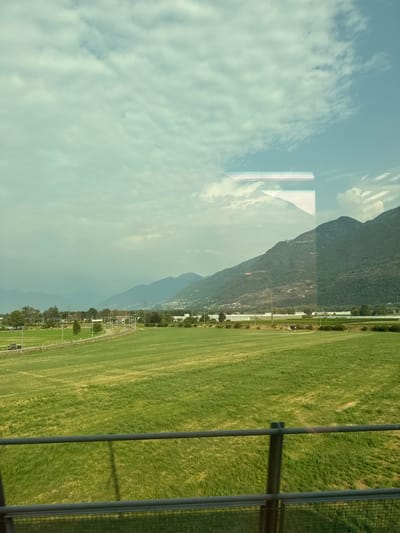 The image captures a landscape view, likely from a moving vehicle, showcasing a vast expanse of green, undulating fields. In the mid-ground, a railway track curves through the fields, with a dark-colored train visible on it. Scattered trees dot the landscape, and a few buildings are nestled in the distance. Beyond the fields and buildings, a range of mountains forms the backdrop, with some of the slopes appearing cultivated or dotted with structures. The sky is partly cloudy, with a hazy quality that suggests either distance or atmospheric conditions. Reflections on what appears to be a window are visible in the upper portion of the image, creating abstract shapes and obscuring parts of the sky and mountains. The scene is predominantly green and blue, with muted tones.