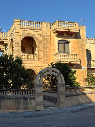 A sunlit, ochre-colored stone building with a clear blue sky dominates the frame, located in Xagħra, Malta. The building features arched balconies with ornate balustrades and stone decorative elements. A low stone wall with a similar balustrade runs along the foreground, encompassing a walkway with stone steps leading up to an archway. Lush green trees partially obscure the lower levels of the building. A street sign is partially visible on the building's facade. The overall scene suggests a residential or possibly a historical property bathed in the warm light of late afternoon.