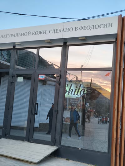 The image shows the entrance to a shop in Орджоникидзевский поселковый совет, Ukraine. The sign above the entrance reads "НАТУРАЛЬНОЙ КОЖИ СДЕЛАНО В ФЕОДОСИИ" (Natural leather made in Feodosia). The shop is currently closed, with the shutters down. Reflections in the glass doors show people walking along the sidewalk outside, including a man in a blue jacket and a group of pedestrians in the distance. A sticker of a red star with a ribbon is visible on the glass. The environment appears to be a street in a tourist area with nearby buildings and mountains in the background.