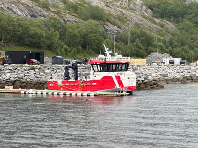 A bright red and white vessel named "NOVA SEA" is docked at a harbor in Stokkvågen, Norway. The boat features a white superstructure with a red hull and a prominent white crane at the bow. The vessel is moored to a wooden dock, which is lined with white and black buoys. In the background, a rocky hillside covered with lush green trees slopes down towards the water. A red car is parked near a black, container-like structure, and a yellow construction vehicle is visible to the left of the container. Piles of rocks and several white, modular buildings are situated on the shore behind the dock. The water is calm and reflects the overcast sky, suggesting a cloudy day. The scene depicts a typical day in a Norwegian coastal town, with maritime activities and land-based infrastructure coexisting. The vessel's name is clearly visible on its stern, and the text "DAMEN Folla" is on its side.