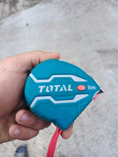 A person's hand is holding a turquoise and white retractable measuring tape with the brand name "TOTAL" prominently displayed. The measuring tape has a red button and indicates "8m". The background appears to be a light-colored, textured surface, possibly concrete or a similar material. The image was taken outdoors. The scene does not contain any specific landmarks or elements that would indicate a location in the city of Kremenets, Ukraine.